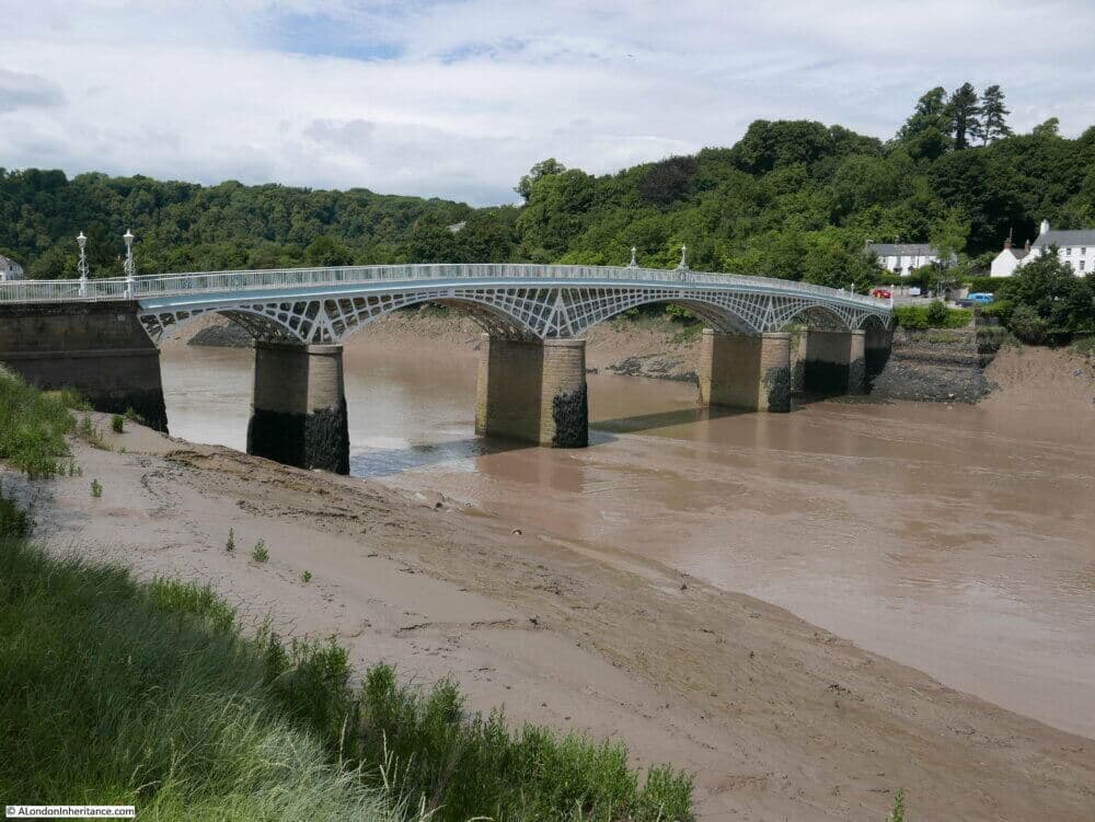 An iron bridge spans over a wide river with muddy banks, surrounded by lush green trees and distant houses. - Home Instead