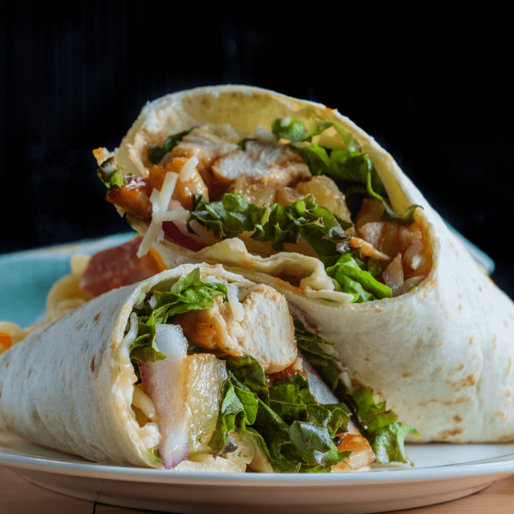 Close-up of a neatly stacked chicken wrap with lettuce, tomatoes, cheese, and onions on a plate. - Home Instead