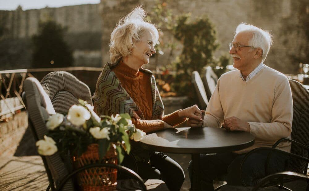 An elderly couple smiles and holds hands while sitting at an outdoor café table with flowers, beneath the sun. - Home Instead