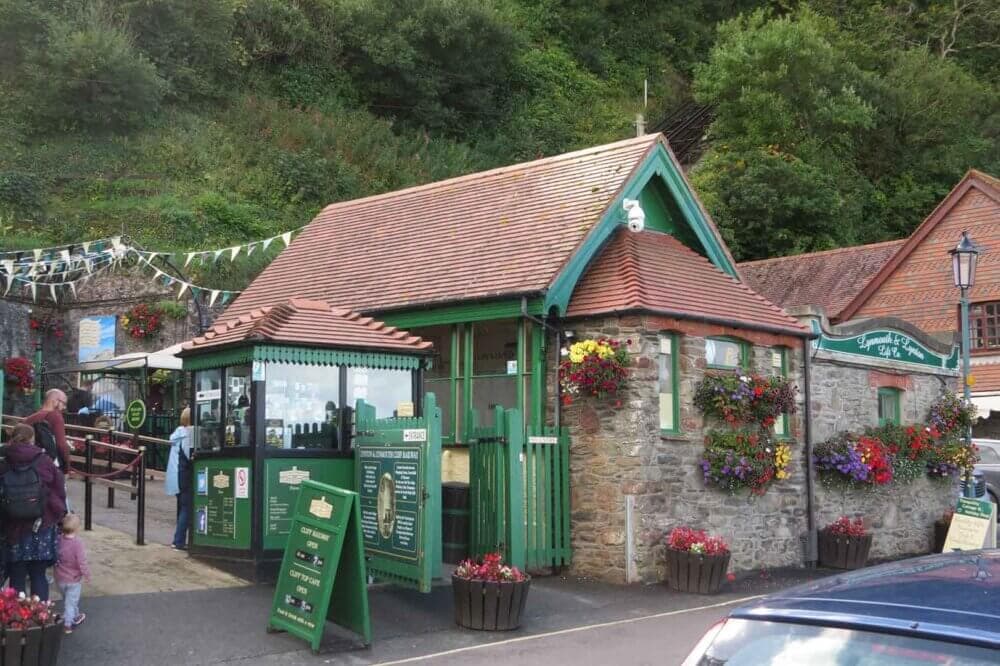 A small, quaint cafe with hanging flower pots, located in a green, hilly area with people waiting outside. - Home Instead