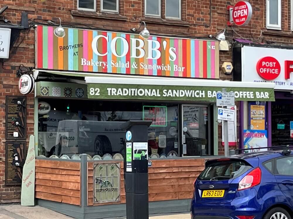 Front view of COBB's Bakery Salad & Sandwich Bar with a colorful striped awning, parked cars, and a Post Office sign nearby. - Home Instead