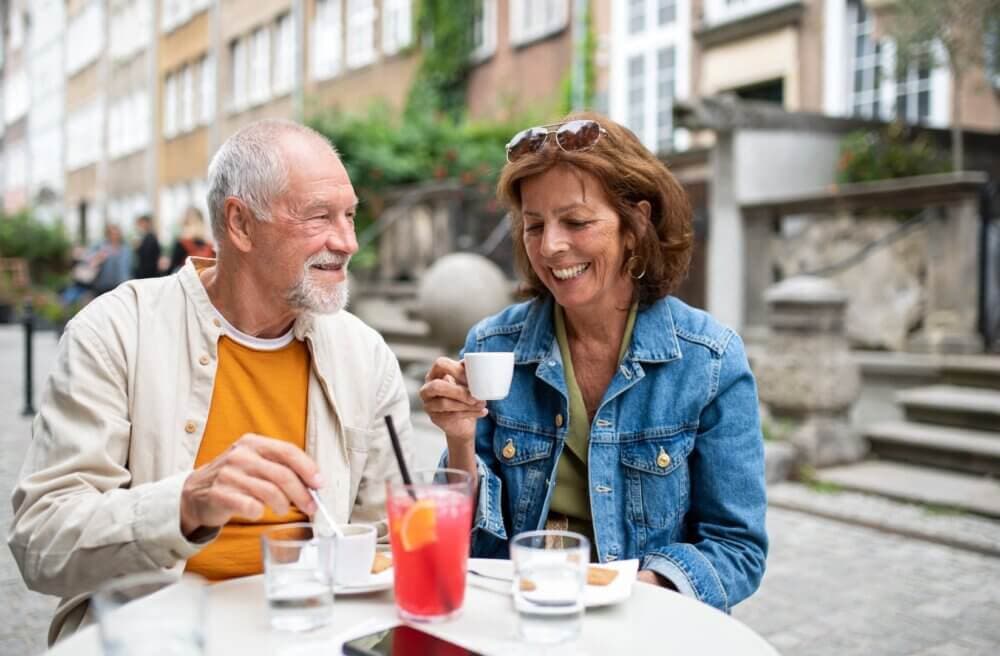 A smiling older couple enjoys coffee at an outdoor café, with drinks and sunglasses on the table. - Home Instead