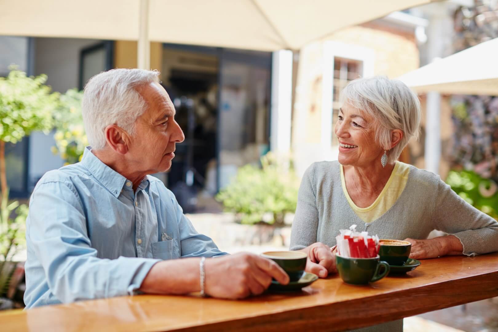 Two elderly people smiling and chatting over coffee at an outdoor café with greenery and umbrellas in the background. - Home Instead