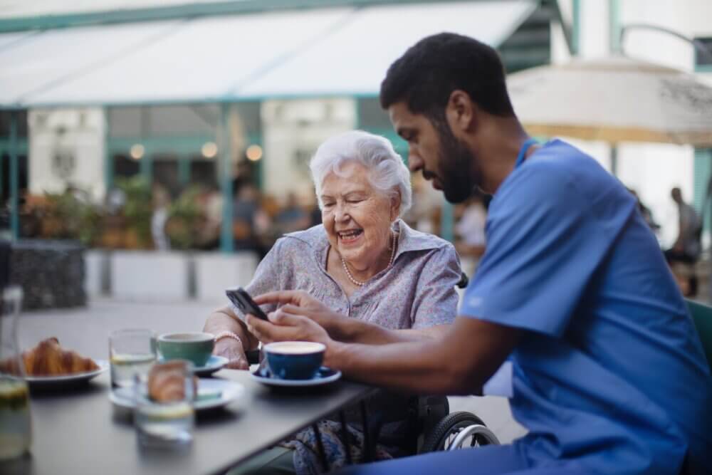 An elderly woman in a wheelchair shares a smile with a nurse while looking at a phone at an outdoor cafe. - Home Instead