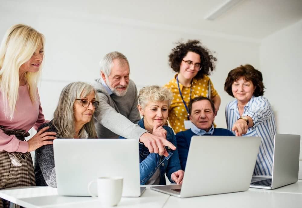 Group of elderly people gathered around laptops, with some pointing at the screens, in a classroom setting. - Home Instead