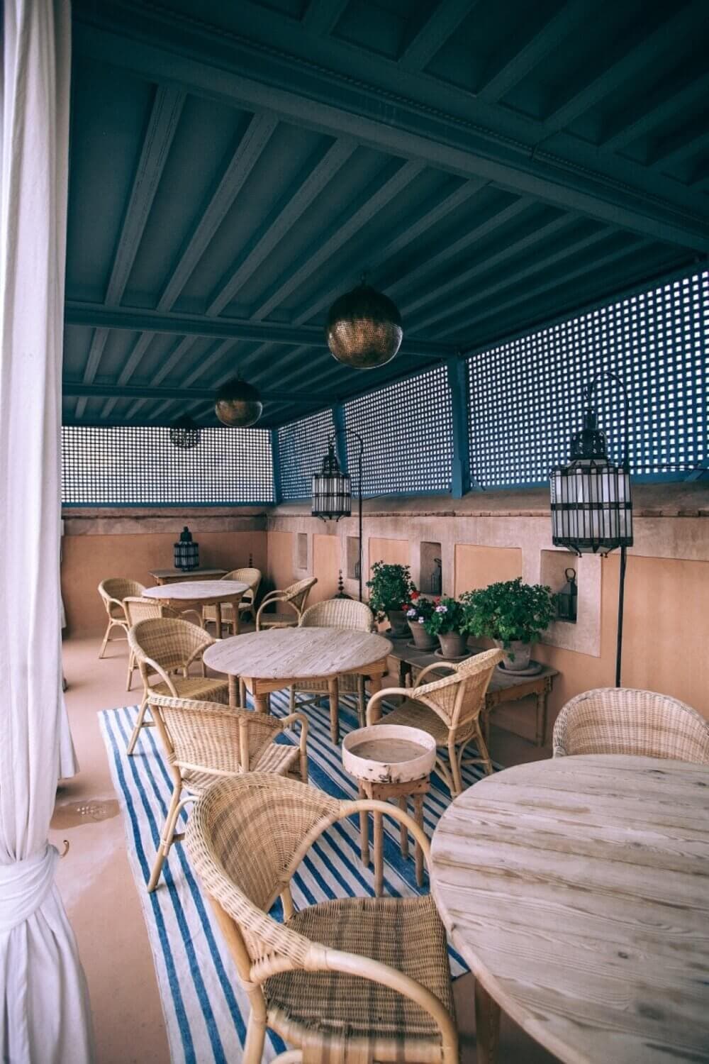 Outdoor patio with wicker chairs, round wooden tables, hanging lanterns, and a striped blue and white rug under a covered roof. - Home Instead