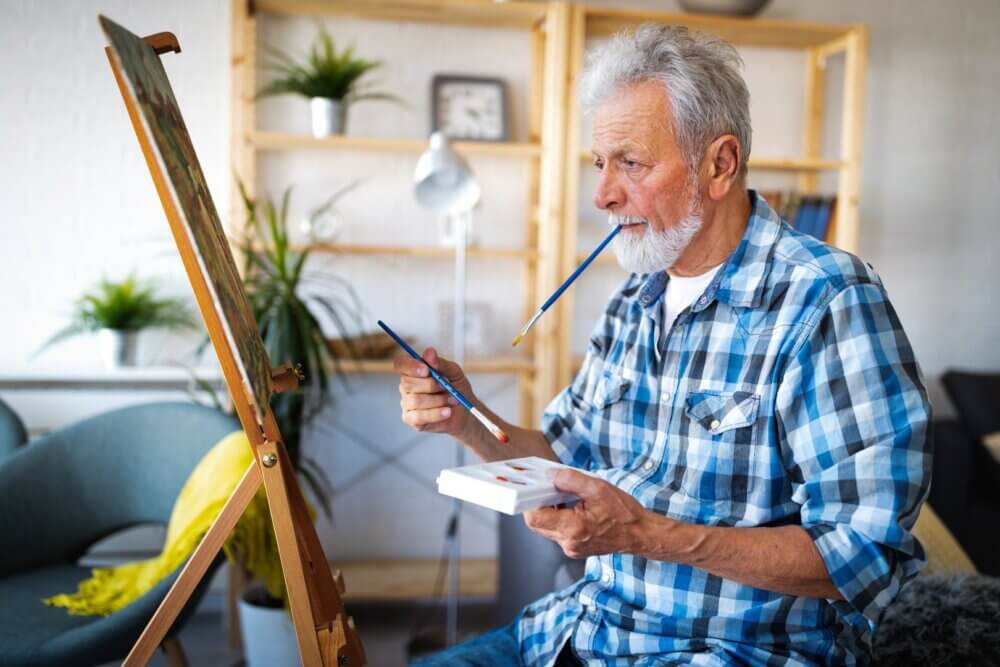 Elderly man painting on a canvas, holding a brush with his mouth and another brush in his hand, in a cosy room. - Home Instead Poole