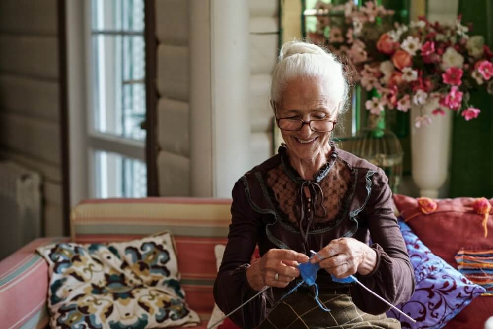 An elderly woman with glasses sits on a sofa, knitting. Bright cushions and flowers adorn the room in the background. - Home Instead