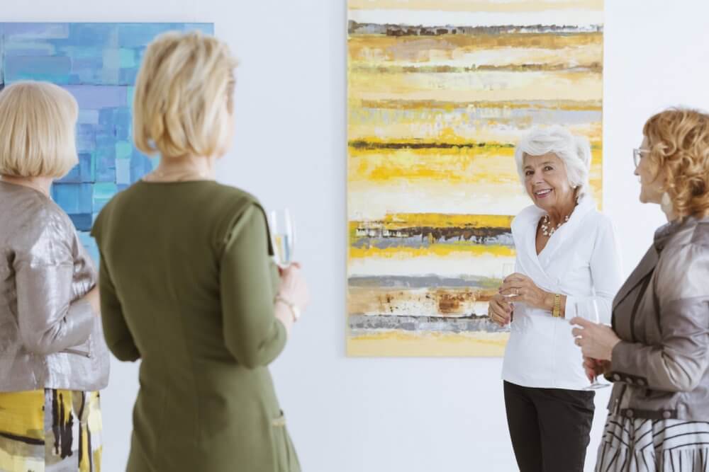 Four women discuss colourful abstract paintings at an art gallery, holding drinks and sharing smiles. - Home Instead Southampton
