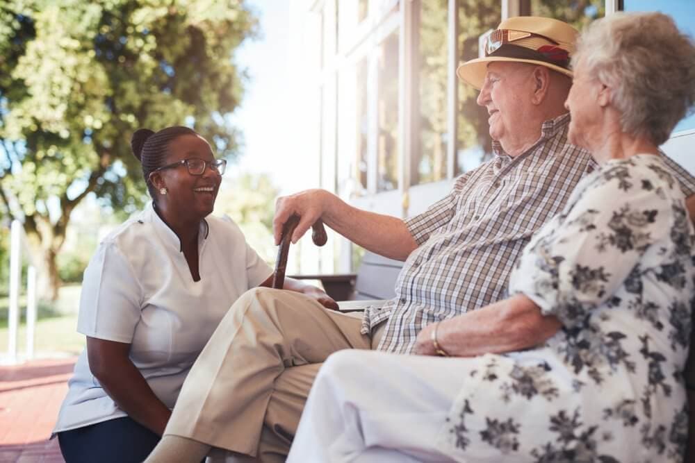 A caregiver smiling and talking with two elderly people sitting outside on a sunny day. - Home Instead