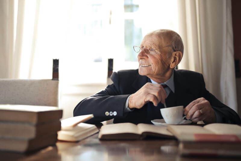 Elderly man in suit smiling, holding a coffee cup, sitting at a table with books in a bright room. - Home Instead
