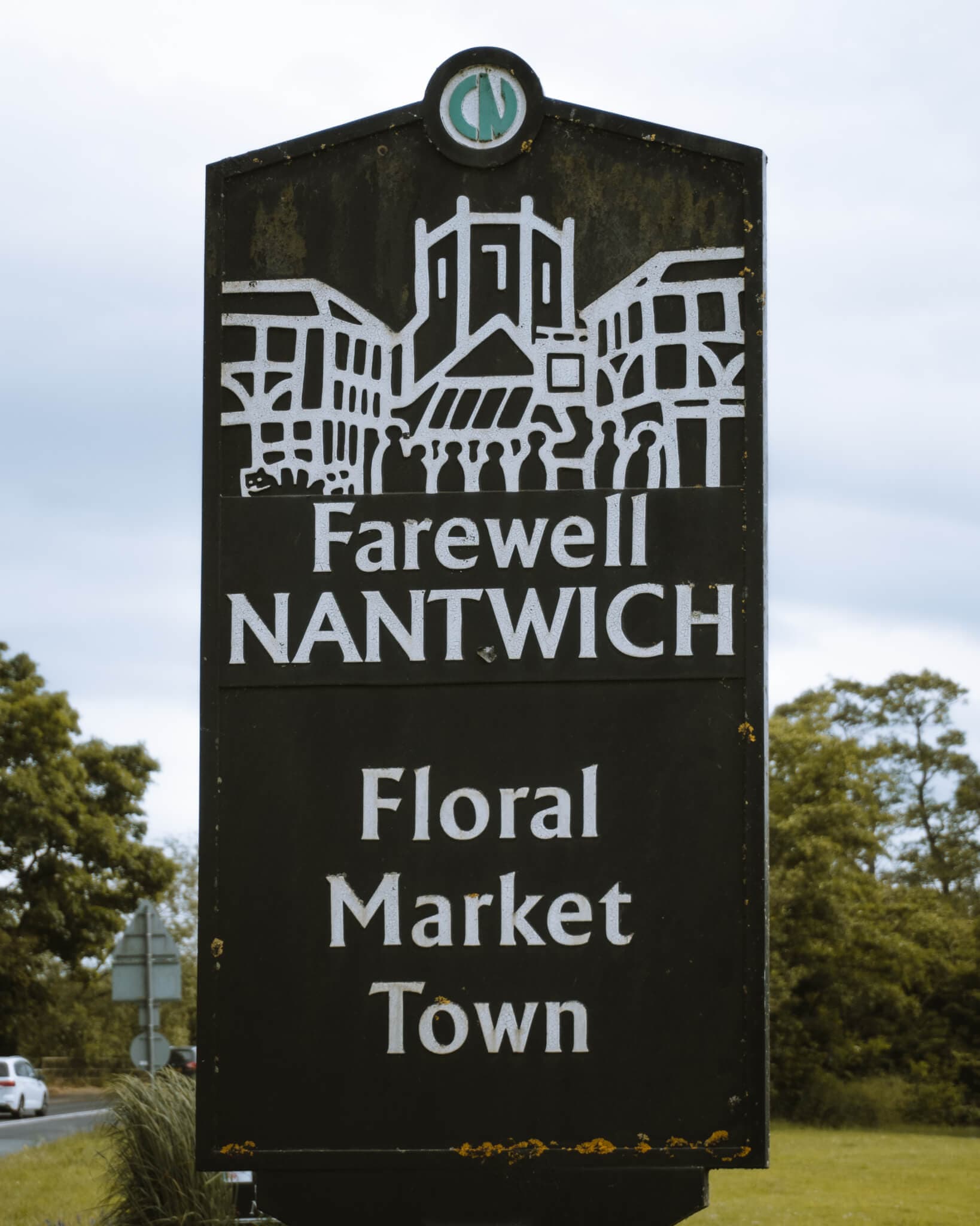 Sign reading "Farewell NANTWICH Floral Market Town" with an illustration of buildings and people, trees in the background. - Home Instead