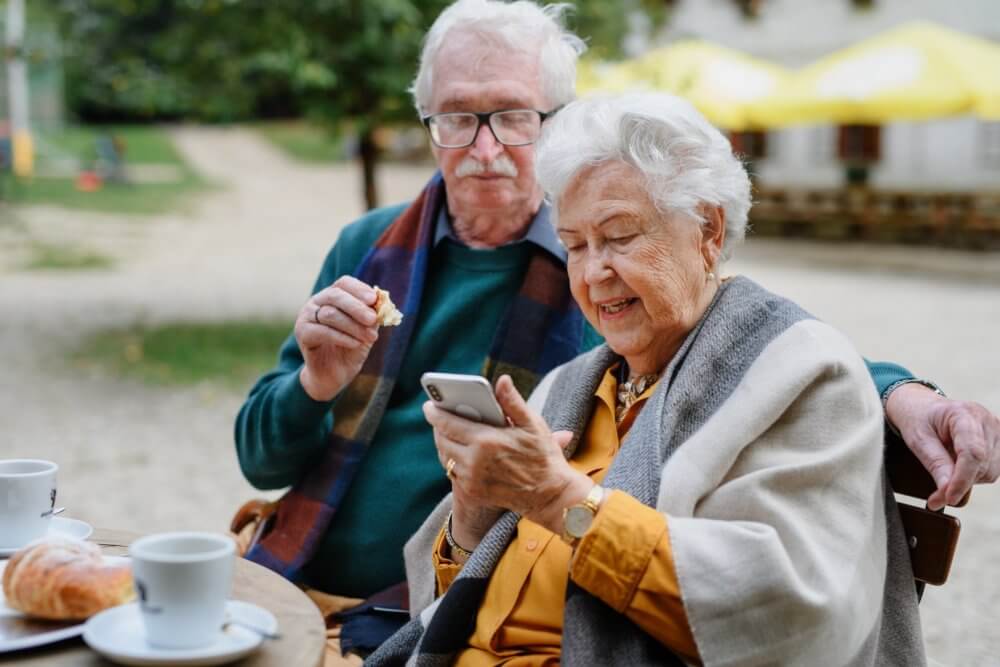 Elderly couple sitting outdoors, drinking coffee and looking at a smartphone together. - Home Instead