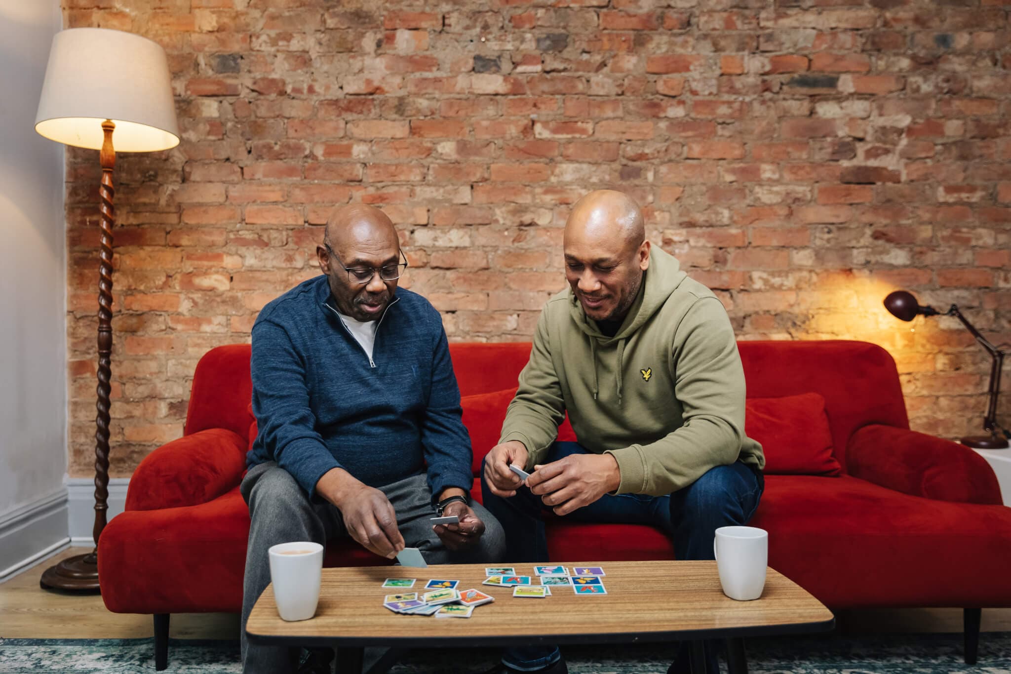 Two men sitting on a red couch, playing a card game at a coffee table, brick wall background, and a floor lamp beside them. - Home Instead