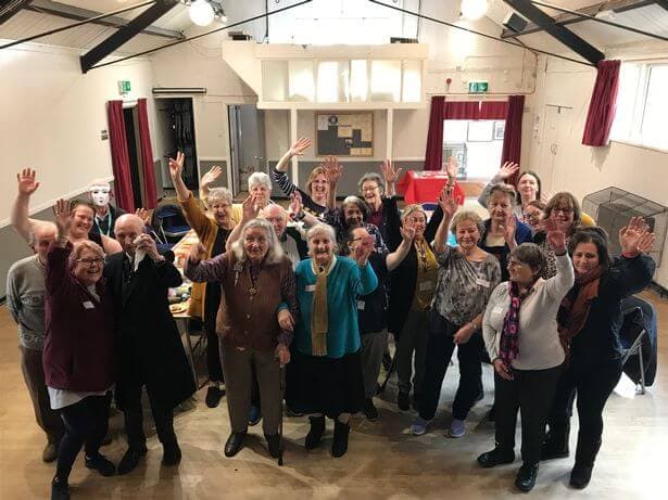 A group of people smiling and raising their hands inside a hall with wooden flooring and high ceilings. - Home Instead