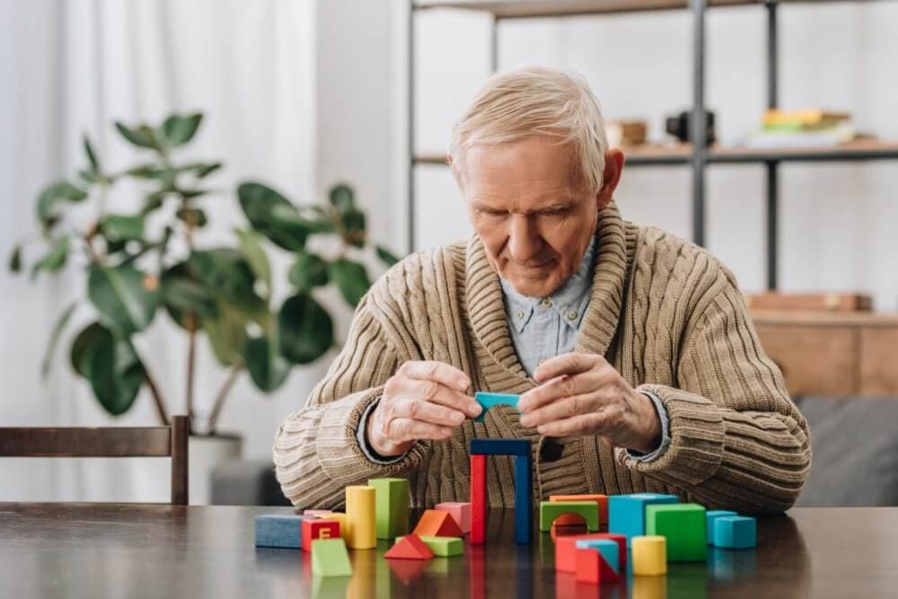 Elderly man in a beige sweater builds a structure with colorful wooden blocks at a table in a cozy room with plants. - Home Instead
