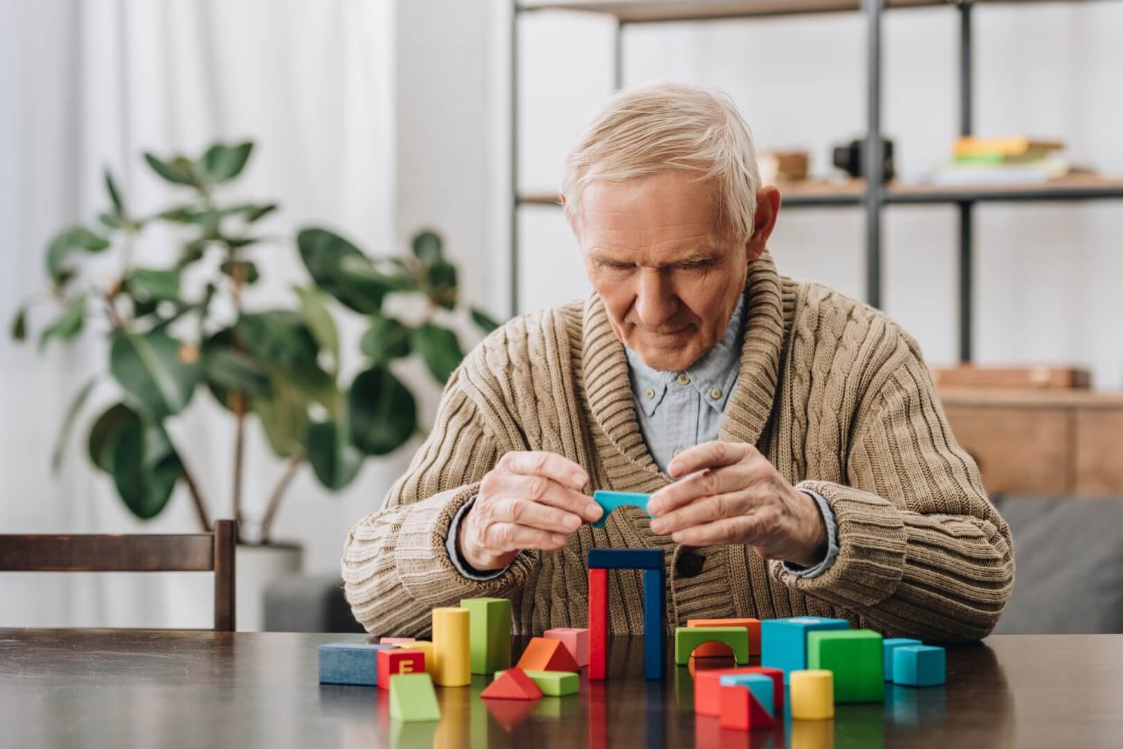 Elderly man in a beige sweater builds a structure with colorful wooden blocks at a table in a cozy room with plants. - Home Instead