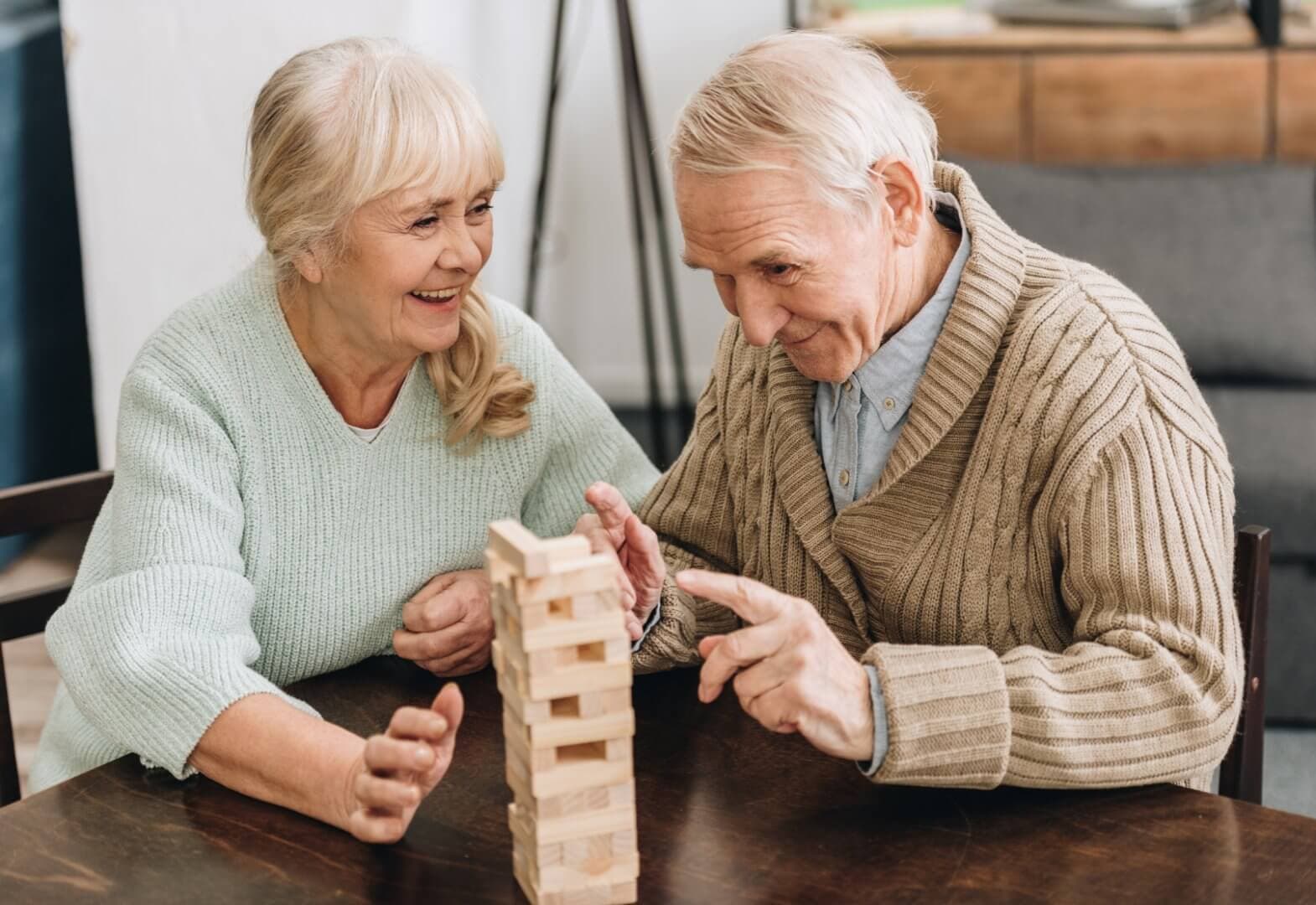 Two elderly people smiling and playing a game of Jenga at a table. - Home Instead