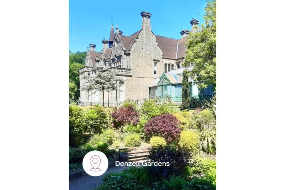 A historic stone building surrounded by lush gardens under a clear blue sky. Text: "Denzell Gardens. - Home Instead