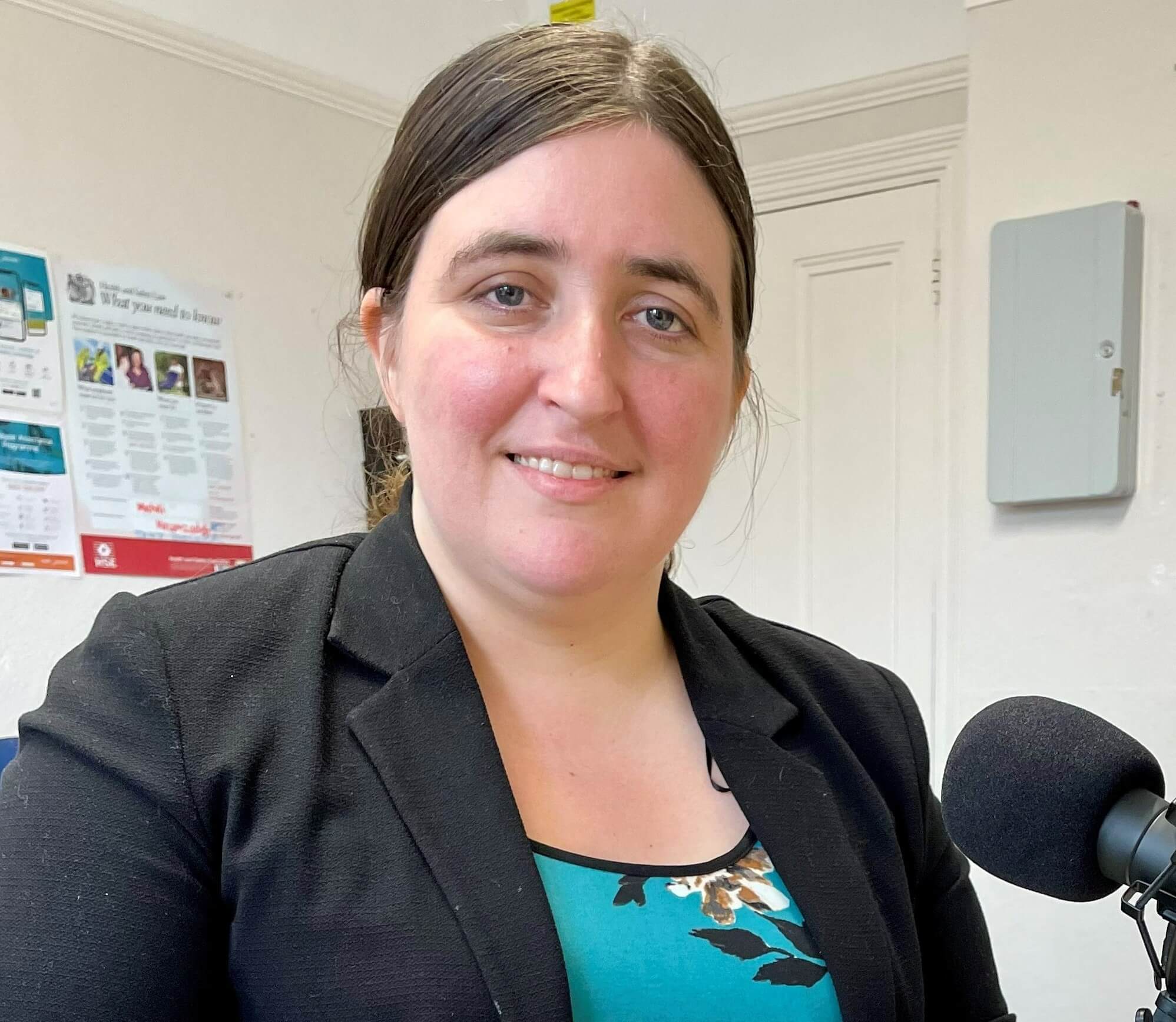 A woman with dark hair is smiling and wearing a blazer; she is sitting in front of a microphone indoors. - Home Instead