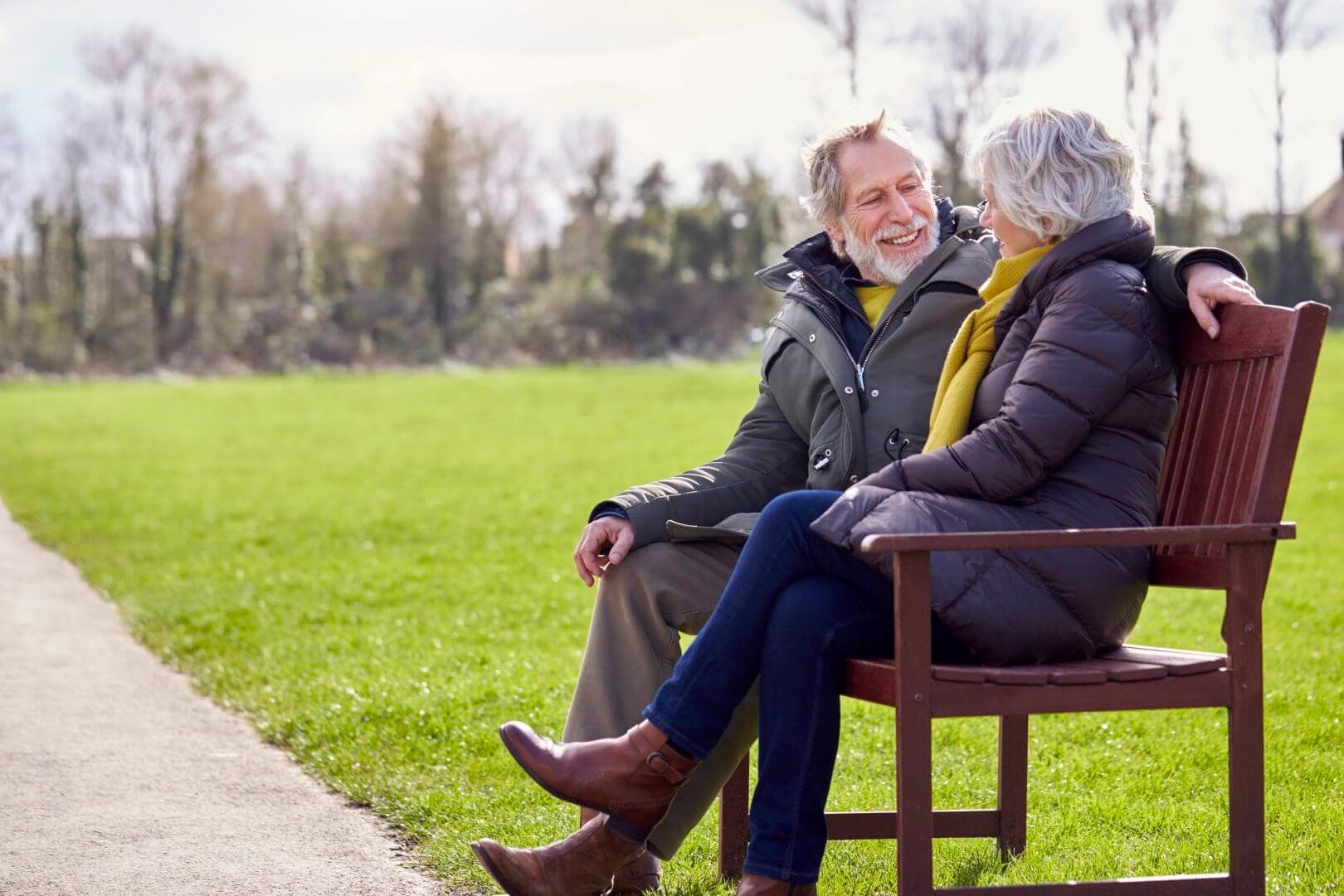 Elderly couple sitting on a bench in a park, smiling and having a conversation on a sunny day. - Home Instead