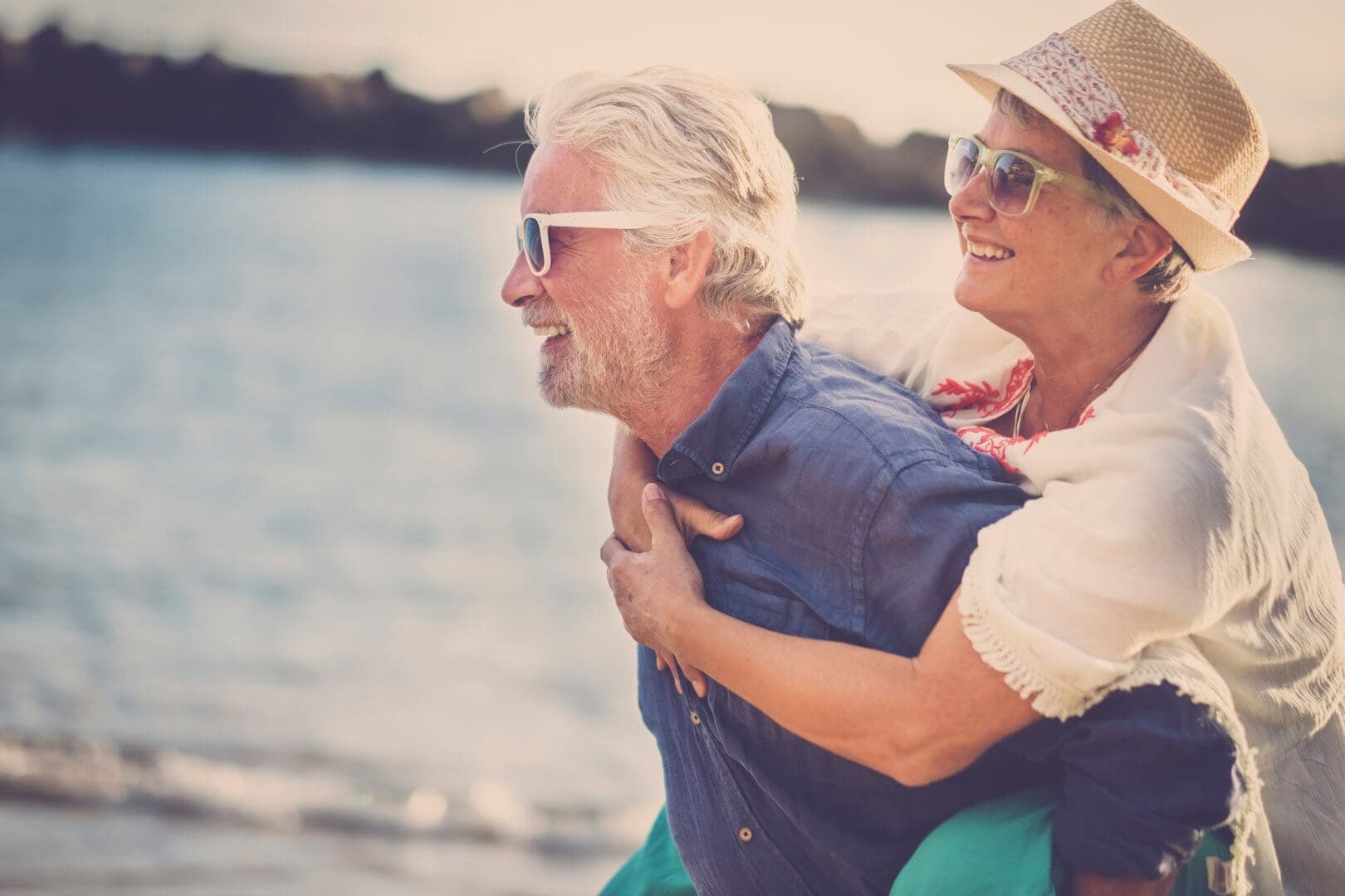 An elderly couple in sunglasses and hats joyfully gives a piggyback ride on a beach with water in the background. - Home Instead