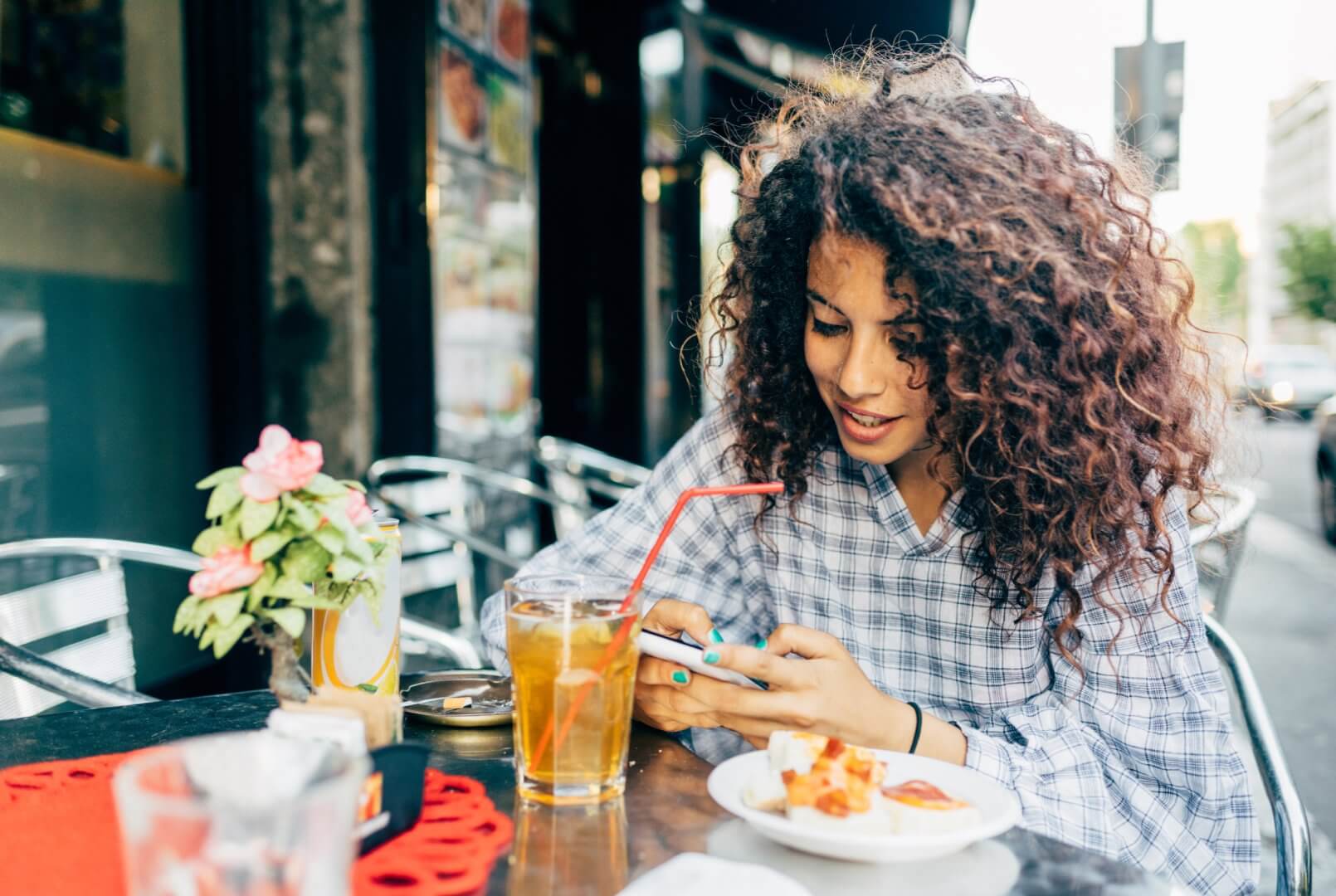 A person with curly hair smiles while looking at their phone in an outdoor cafe, with a drink and food in front of them. - Home Instead