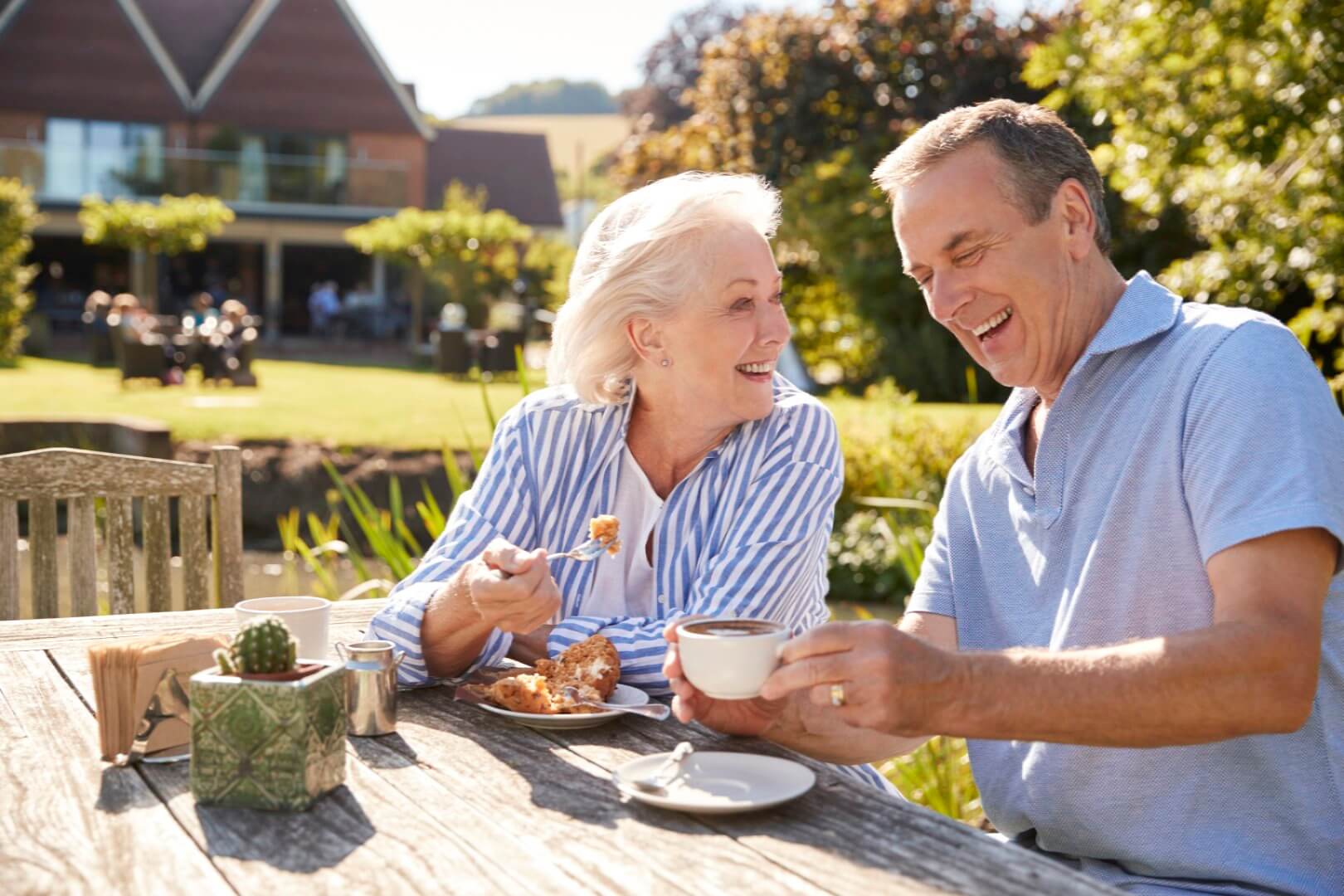Senior couple laughing and enjoying a meal together outdoors at a wooden table in a garden setting. - Home Instead