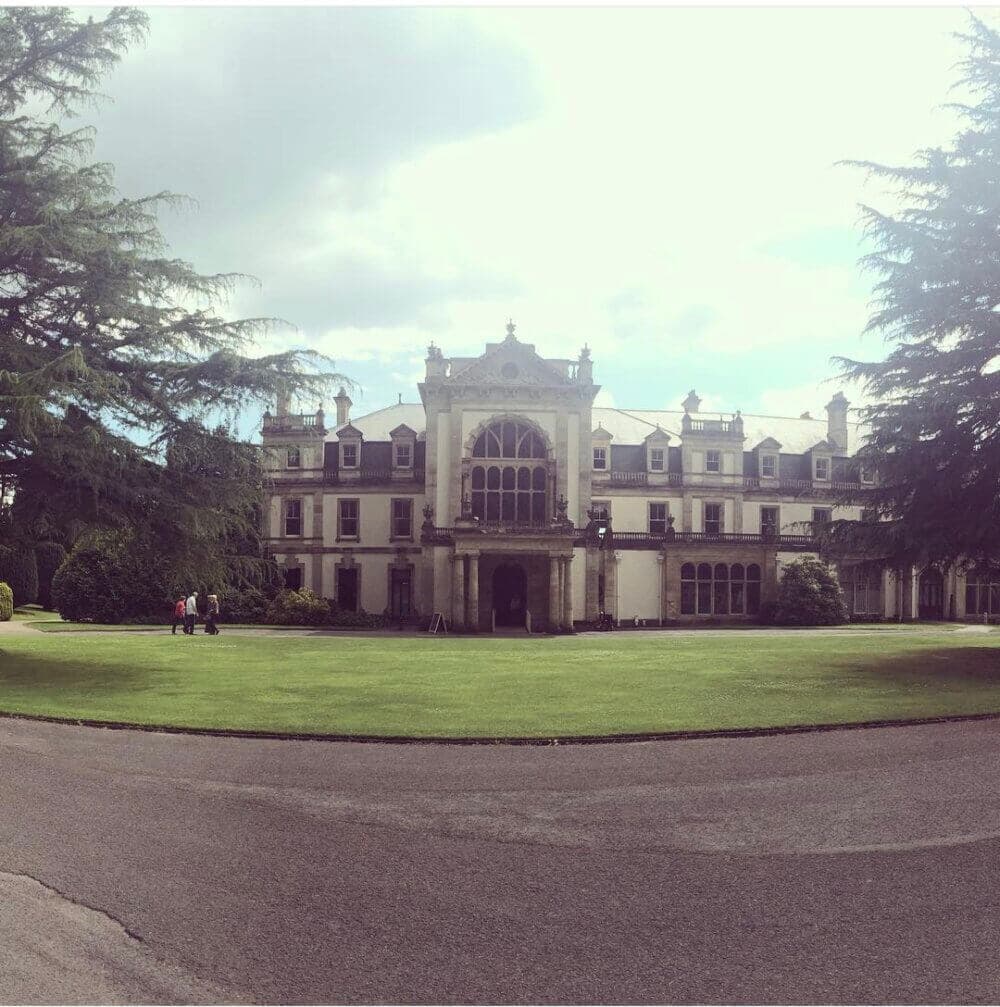 A large historic mansion with a well-manicured lawn and trees, against a partly cloudy sky. - Home Instead