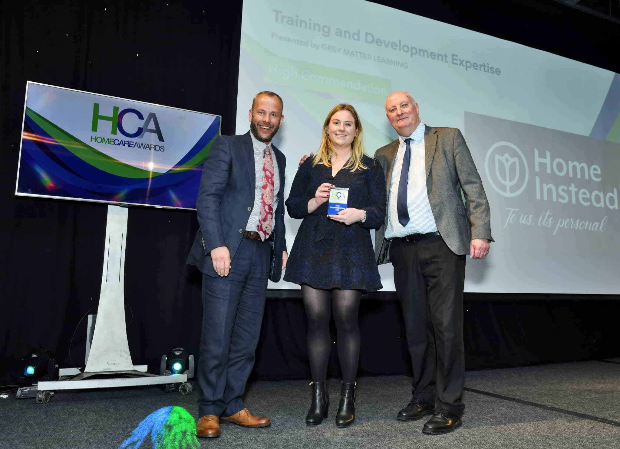 Three people standing on stage at an awards event with a "High Commendation" certificate, "Home Instead" in the background. - Home Instead