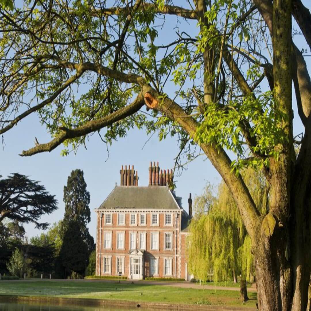 A large historic brick house framed by trees, with lush greenery and clear blue skies in the background. - Home Instead