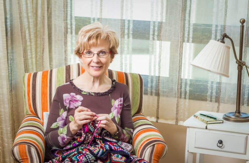 Woman smiling while knitting in a cozy chair beside a lamp and table. - Home Instead