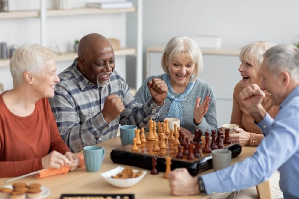 Group of smiling seniors playing chess at a table, celebrating a move together with drinks and snacks around. - Home Instead