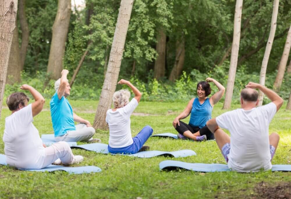 A group of people doing seated exercises on mats in a park, led by an instructor. - Home Instead