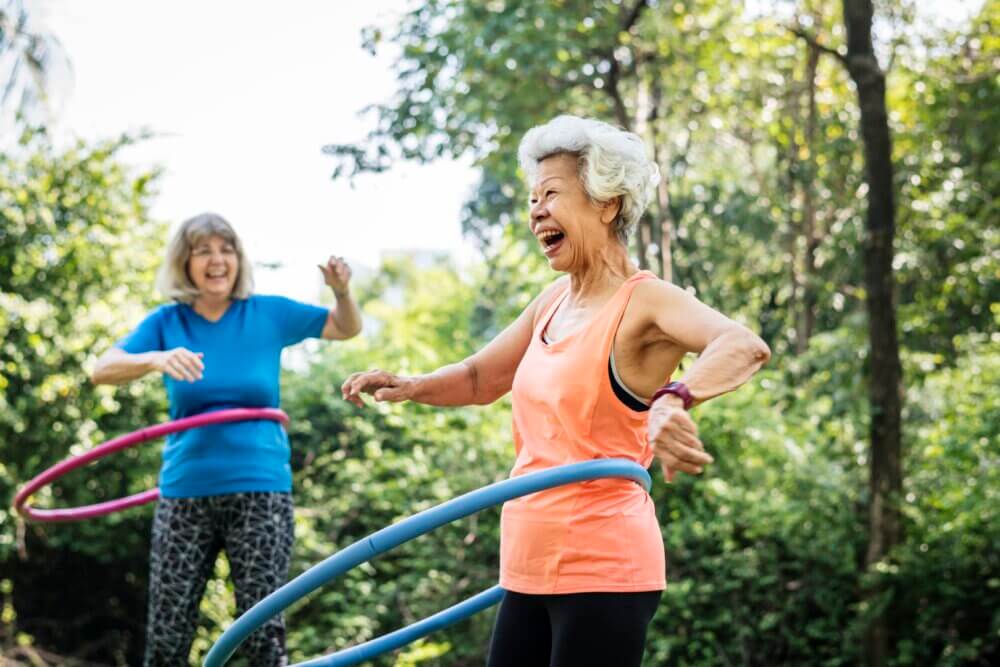 Two smiling older women hula-hooping in a green outdoor setting, one in a blue top and the other in an orange top. - Home Instead Poole