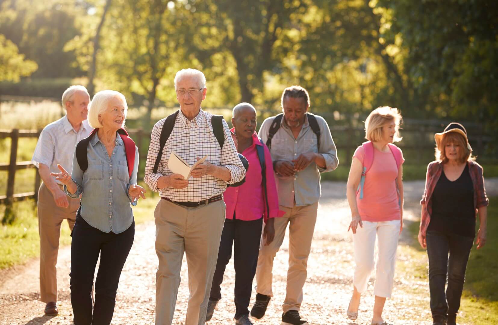 A group of older adults walking outdoors on a sunny day, wearing casual clothing and carrying backpacks. - Home Instead