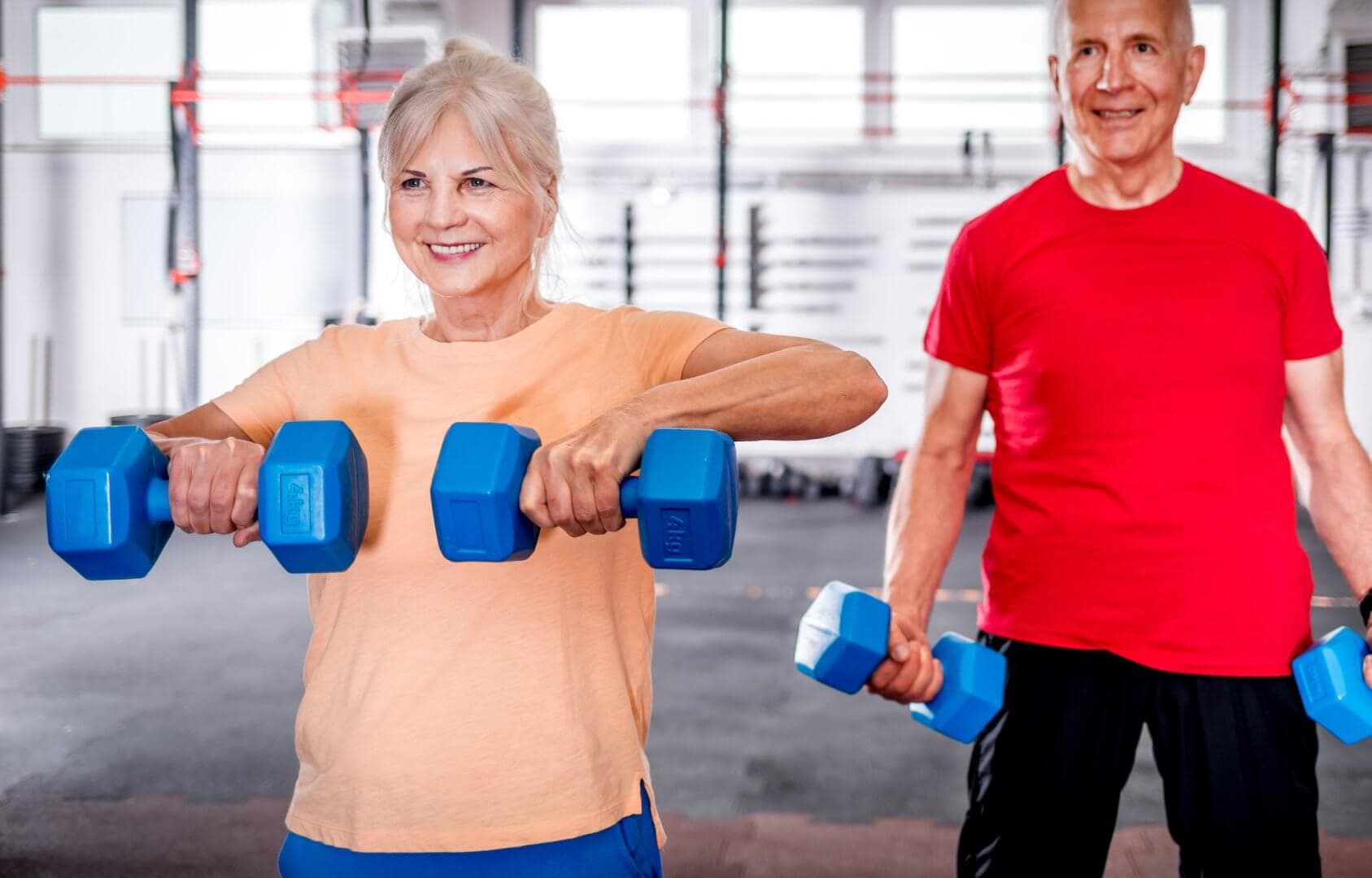 Two older adults lifting blue dumbbells in a gym, smiling. The woman wears orange, the man wears red. - Home Instead