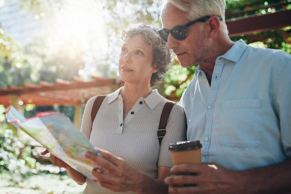 Older couple studying a map outdoors, with the man holding a coffee cup and both dressed casually under the sunlight. - Home Instead