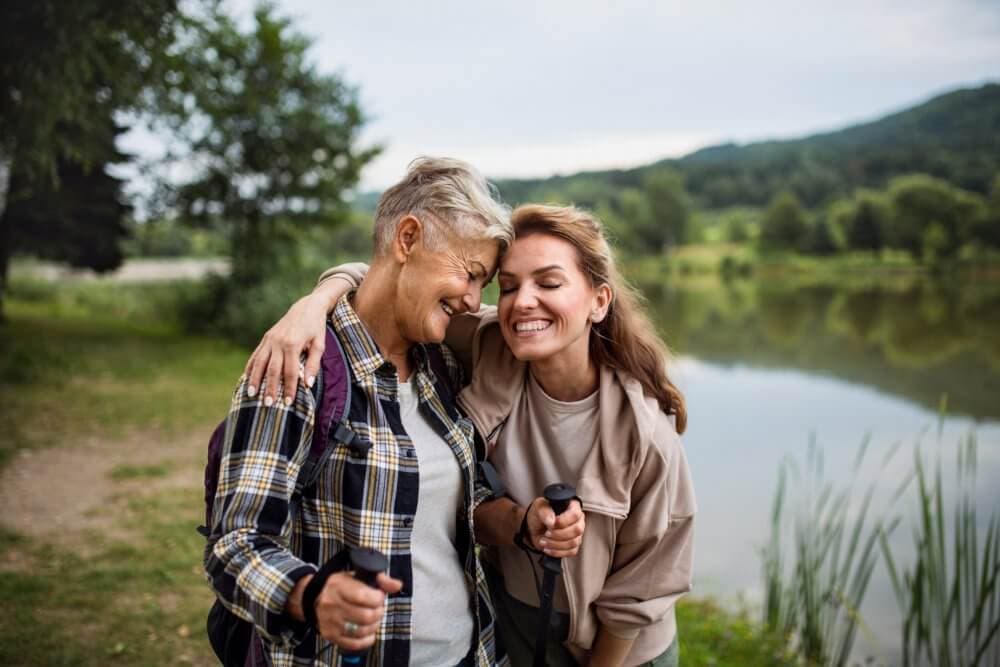 Two women smiling and embracing each other while holding walking sticks near a lake with green hills in the background. - Home Instead