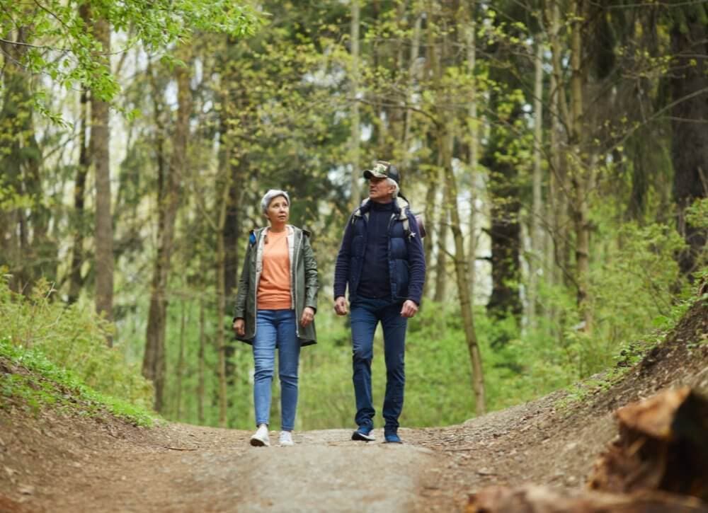 Two people in outdoor gear walking on a wooded trail, surrounded by lush green trees and foliage. - Home Instead