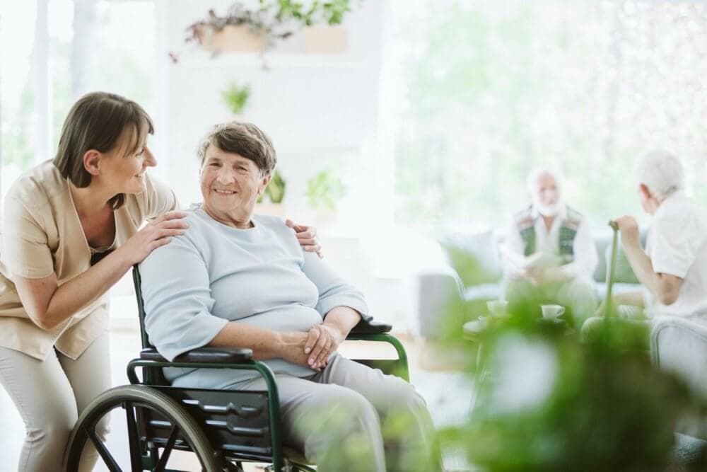 A smiling elderly woman in a wheelchair with a caregiver, with two other elderly people in the background in a bright room. - Home Instead Southampton