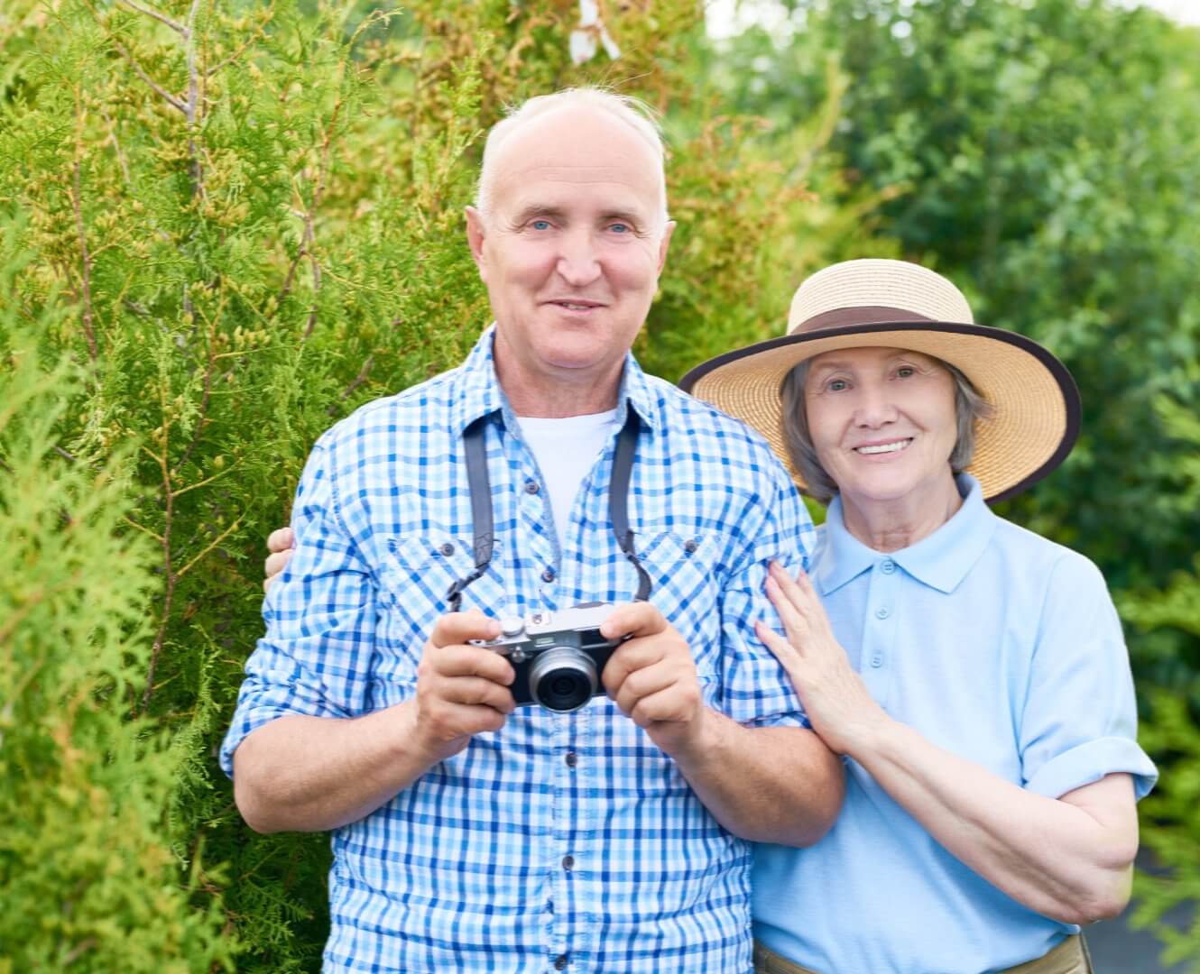 Elderly couple smiling outdoors; man holding camera and woman wearing a sun hat, standing by greenery. - Home Instead