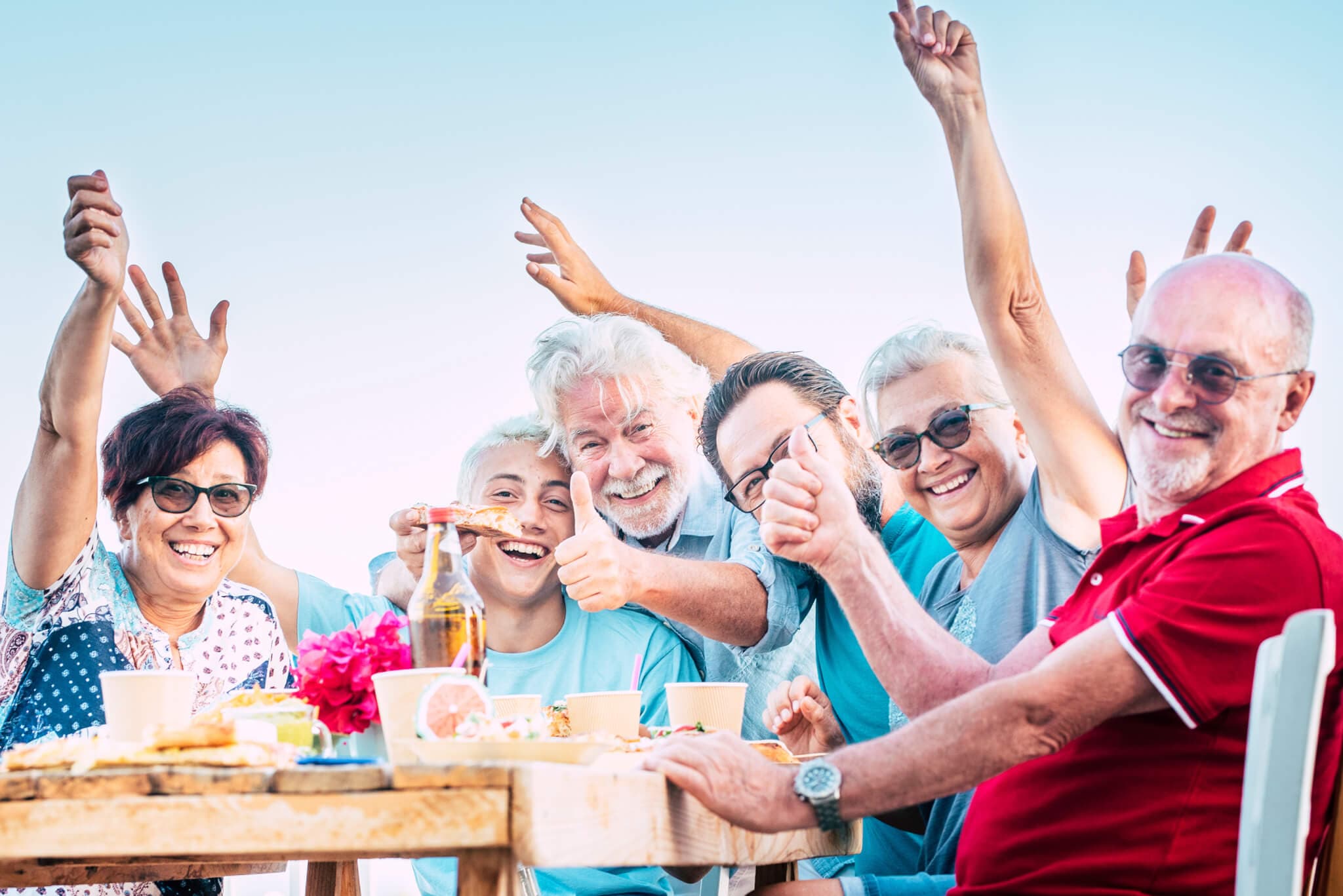 A group of older adults at an outdoor table smiling, raising their hands, and enjoying food and drinks together. - Home Instead
