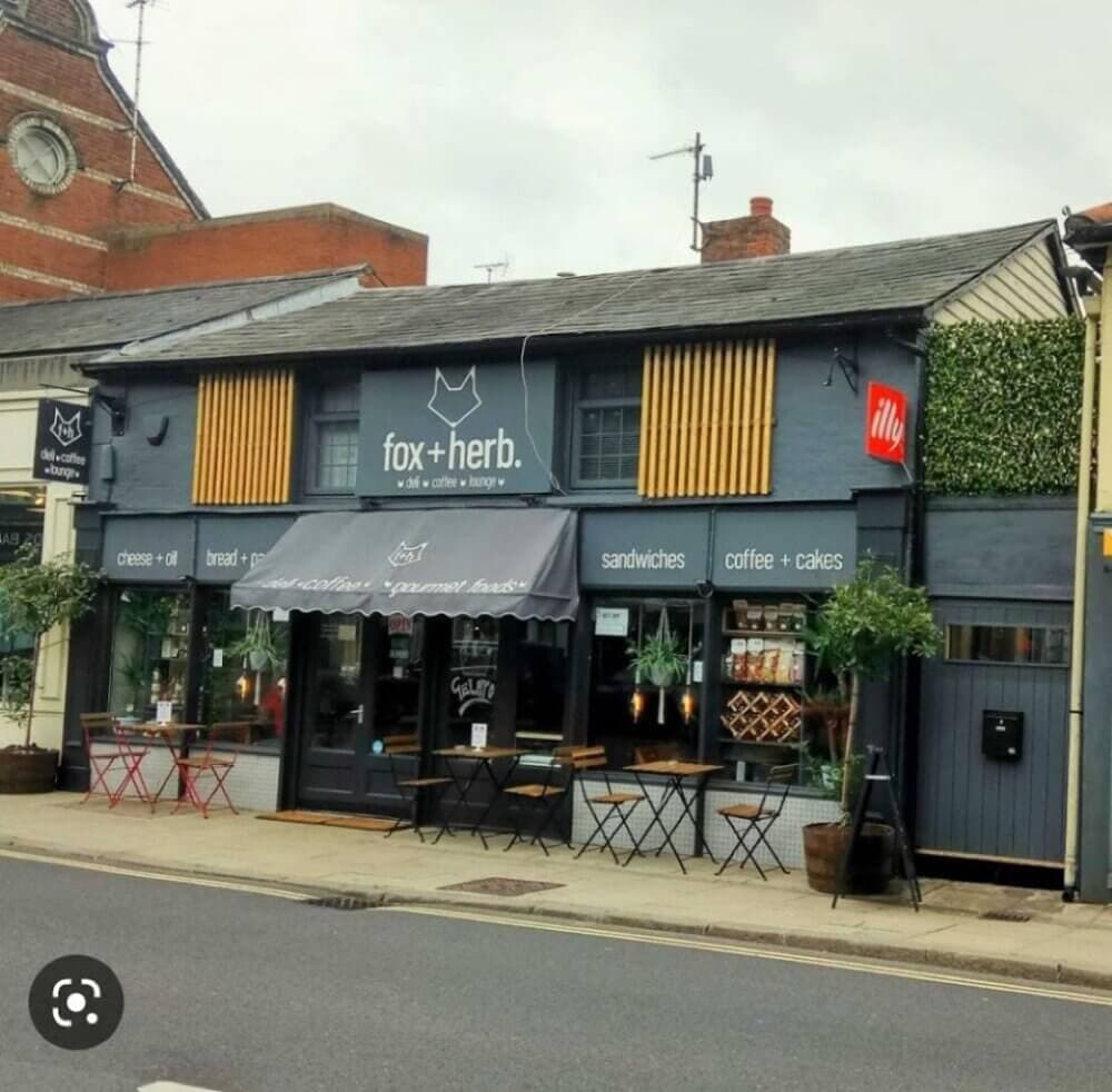 A cozy cafe named "fox+herb" with outdoor seating and a modern grey exterior, featuring greenery and a chalkboard sign. - Home Instead