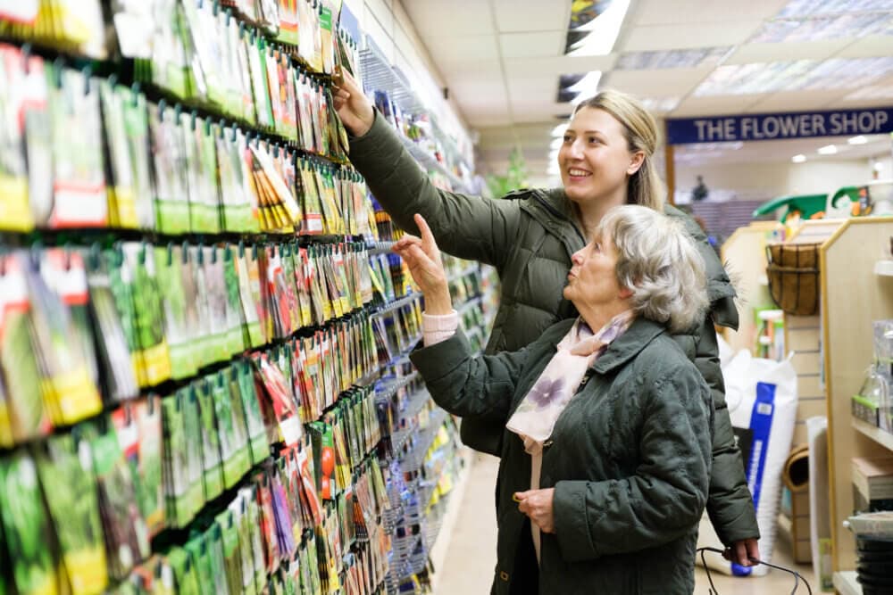 Two women in a store looking at packets on a shelf, one reaching up and the other pointing to an item. - Home Instead