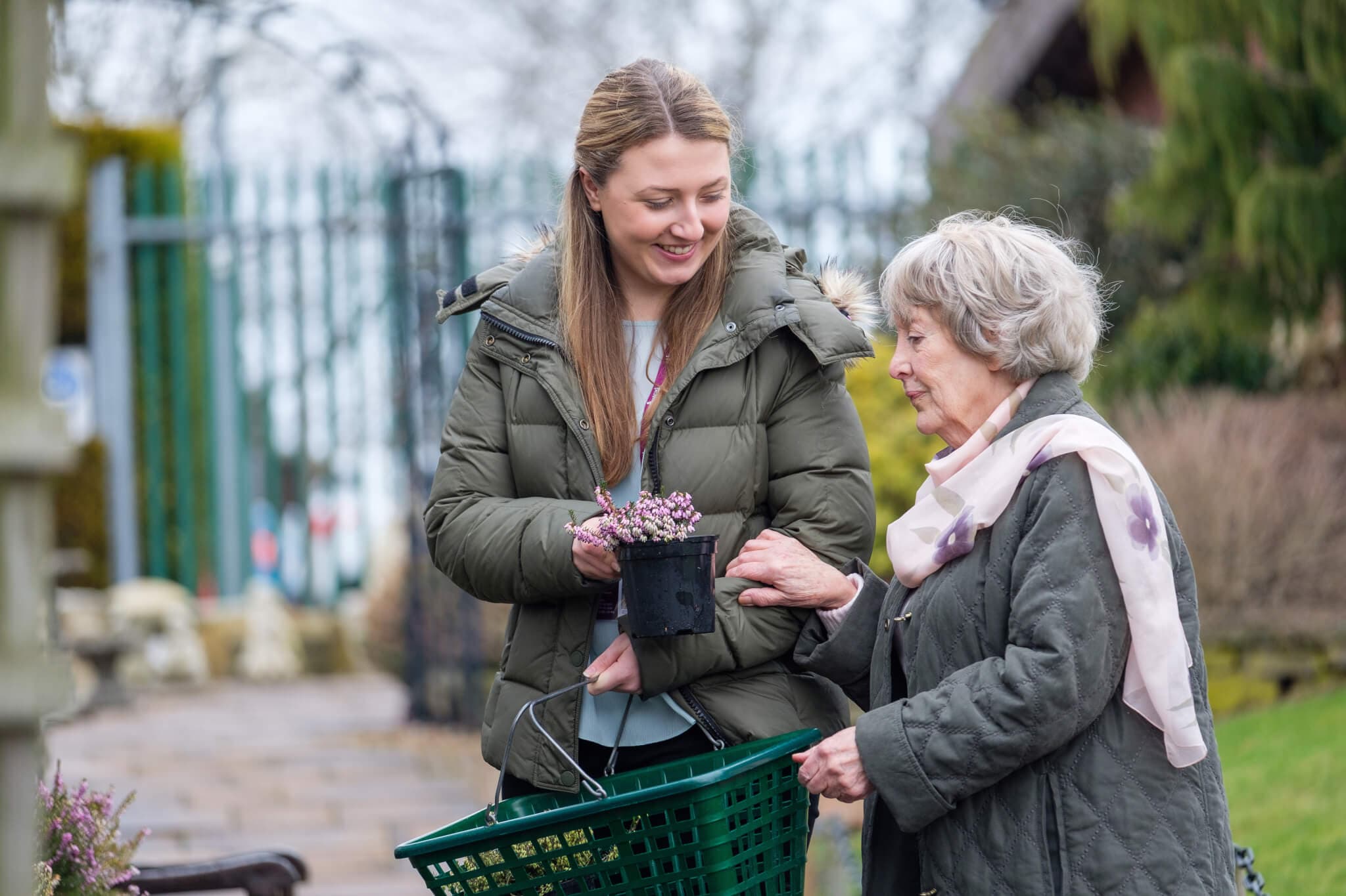 A young woman helps an elderly woman hold a plant pot while they are shopping outside. - Home Instead
