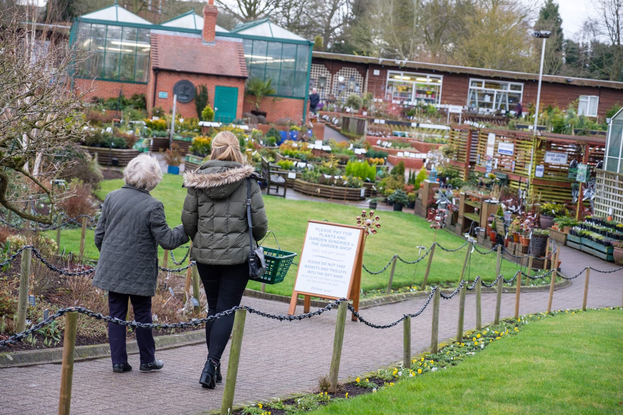 Two women walking towards a garden center surrounded by plants and flowers. A sign and greenhouse visible ahead. - Home Instead