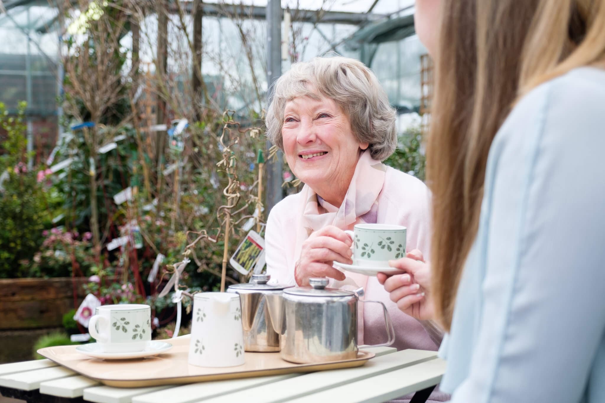 Elderly woman smiling and holding a teacup, having tea with a younger woman in an outdoor garden setting. - Home Instead
