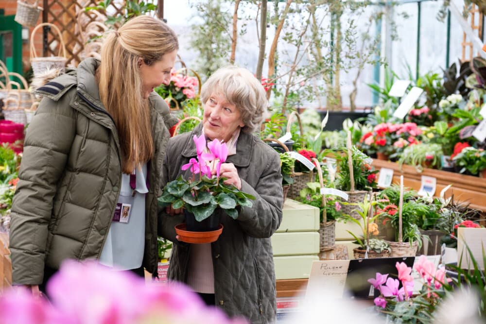 Two women smiling and holding a potted plant in a garden center filled with various flowers and plants. - Home Instead
