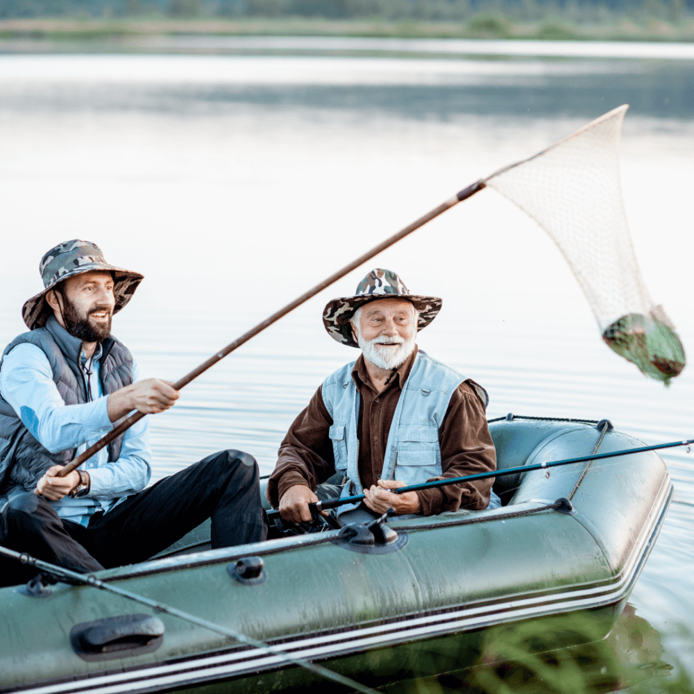 Two men in fishing hats are in a boat on a lake, lifting a fish with a net. - Home Instead